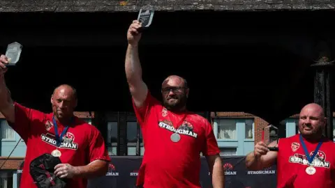 James Merry Three men in red t-shirts stand with hands in the air holding small glass trophies.