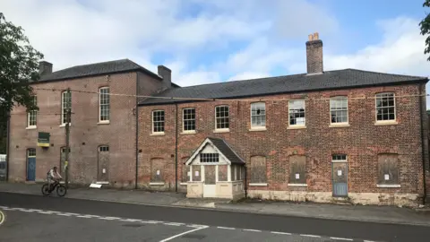 LDRS The building viewed from the other side of the road reveals it is on a hill. The ground floor windows are boarded up but the upper floor windows are exposed. There is a white timber porch with a pitched roof on the main entrance on to the pavement.