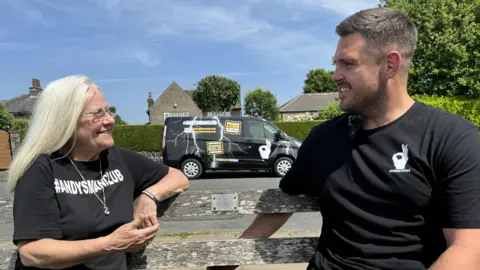 A older woman with shoulder-length white hair and glasses sits on a bench next to a man with short brown hair and brown stubble. They both wear black T-shirts with white lettering that reads: Andy's Man Club and It's Okay to Talk.