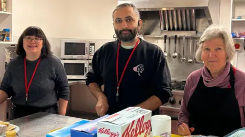 King's Arms Project Three people, a woman, a man, and another woman, standing in a kitchen They all have red lanyards on. The woman on the right has dark shoulder length hair, with glasses and is wearing a dark jumper, the man in the middle has a dark jumper on and a woman on the right is wearing a black apron with a purple jumper underneath. They are in a kitchen, with a microwave, utensils and a cooker behind them. They are by a silver bench with food on it. 