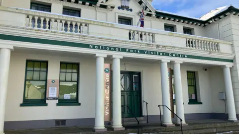 The exterior of a victorian style building, with stone columns along the front and large doors which are painted green.  A sign above the entrance reads National Park Visitor Centre. 