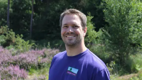 Ashdown Forest A smiling man in a purple T-shirt stands in front of heather and green forest