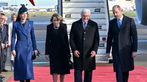 Reuters Four people walk on red carpet in front of a plane with Catherine wearing a blue jacket, and Elke Budenbender wearing a black dress, Frank-Walter Steinmeier wearing a black suit jacket and William wearing a black suit jacket