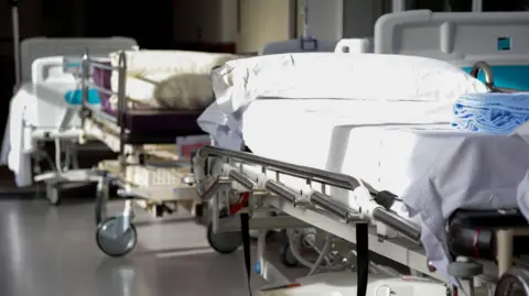 Two hospital beds are seen with white sheets, pillows and a blue blanket on the end of one of them. The sun is shining through an unseen window on to the beds.