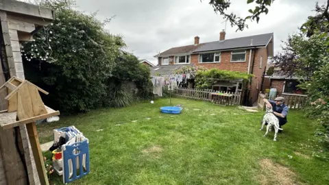 A semi-detached house and its grass garden. Features of the garden include a whirly and a wooden building. Peter Chapman is petting a large dalmation.