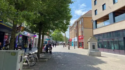 A picture of Staines high street, with trees, benches, bike railings, people and shops.