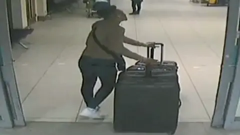 National Crime Agency CCTV footage of a man with a beige jumper on, dark trousers and white trainers entering a door wheeling three large black suitcases
