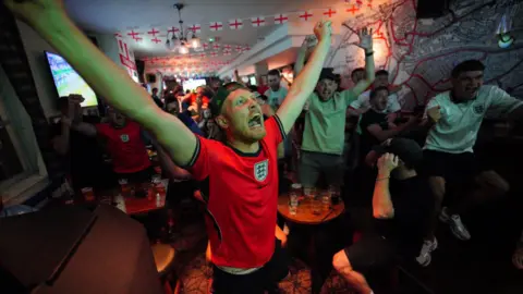 A close-up of a fan wearing a red England shirt, with his arms outstretched in the air and his mouth open with joy. Other fans are celebrating similarly in the background of the pub room, which has England flag banners strung from the ceiling 