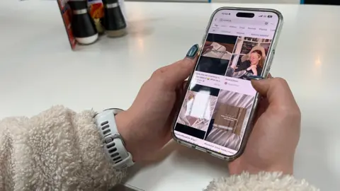 A woman with manicured nails holding a phone in a cafe. The screen shows images of people and books. 