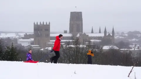 PA Media A young boy in a red coat pulls a girl sitting on a red sleigh on a hill in snow. In the background a cathedral can be seen.