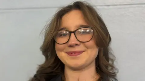 Claire Hamilton/BBC Lily Shirley has brown hair an large black spectacles.