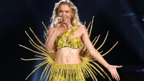 Zara Larsson wearing a yellow sparkly two piece outfit. She has long blonde hair and she is singing into a microphone.