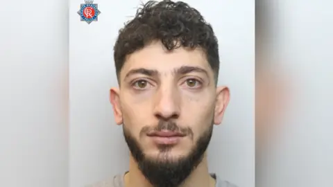 A police mugshot of a man looking at the camera. He is expressionless and has a dark hair, a dark beard and moustache.