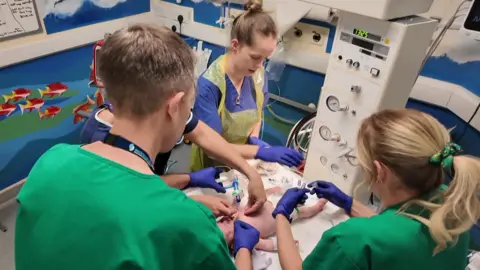 The Southampton Oxford Retrieval Team caring for a baby in hospital in Winchester. There are four people standing around the bed with a small child. Two people are wear blue scrubs, the other two green. The walls of the room are blue with a painted fish mural.