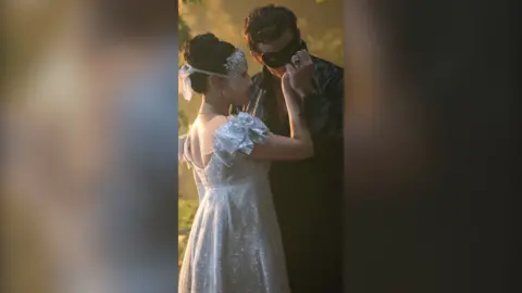 Benedict Bridgerton in black clothes, kissing the hand of Sophie Baek, in a white dress. Both are masked.
