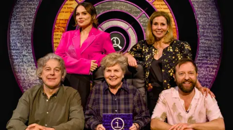 Sandi Toksvig sits in the centre. Surrounding her are Alan Davies, Joanne McNally, Sally Phillips and Andrew Maxwell