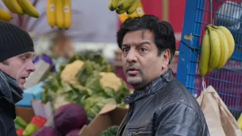 BBC/Keiron McCarron Nitin is wearing a black leather jacket and in the Albert Square Market on the set of Eastenders