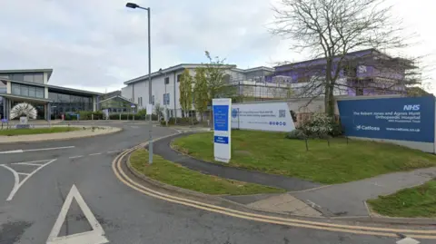 The outside of a hospital building - on the left, there is a glass-fronted building with a large sculpture outside it that looks like a dandelion. In front of the sculpture is a roundabout. There are more hospital building to the right, and fencing and green grass with a blue sign that reads "NHS, The Robert Jones and Agnes Hunt Orthopaedic Hospital"