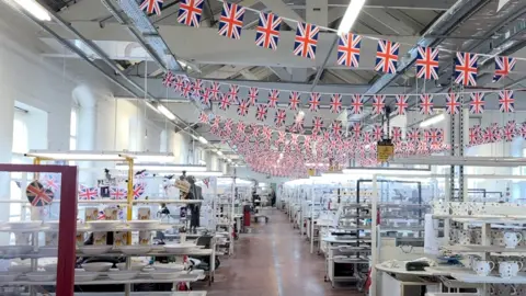 A factory showroom festooned with union jack bunting and containing rows of shelves showcasing the company's pottery in a variety of designs.
