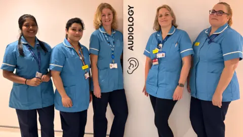 NHS Somerset FT A group of five female clinicians in a uniform of short-sleeved pale blue tops and dark blue trousers, all smiling to camera standing by a sign saying 'audiology' in a hospital corridor