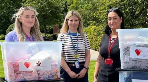 Three women - including Gemma Lowery on the right, who has long dark hair and is wearing black - standing behind piled-up plastic tubs, that are filled with everyday essentials.