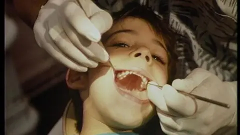 A dentist examines a child's teeth holding metal instruments in the child's mouth.