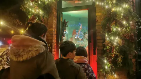 OUTSIDE Three people stood in front of an open door with musicians stood inside the house which is covered in Christmas lights. Their backs are to the camera and they include a woman in a hooded coat with dark hair and two children in front of her with dark hair.