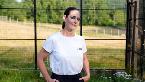 PA Media Kirsty Gallacher wearing a white t-shirt with her black hair tied back. She is stood in front of a metal enclosure.
