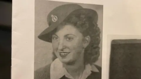 Dorrie Pack The image is a photograph of a leaflet showing Dorrie as a young woman in her Land Army uniform in a black and white picture. She is looking off to the left of the camera lens. She has dark, curly hair and dark lipstick. 
