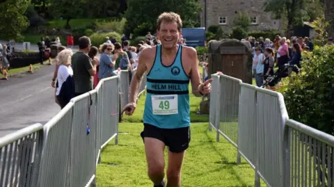 David Griffin, a 66-year-old man, is running towards the camera. He is wearing a blue vest which has the number 49 on it and is running between silver metal barriers which suggest the end of a race. In the distance a number of people are watching the runners and applauding.