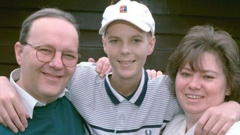 An old picture of a young Christopher Lucas standing with his arms around his parents' necks