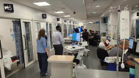 University Hospitals Birmingham A busy hospital ward filled with staff and patients, there are patient bays running down the left side of the ward and to the right staff are seen working on computers