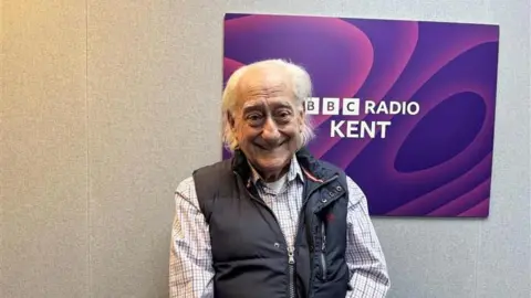 Jack Valpy/BBC A man with grey hair, standing in front of a purple BBC Radio Kent sign. He has a black coat and chequered shirt on.