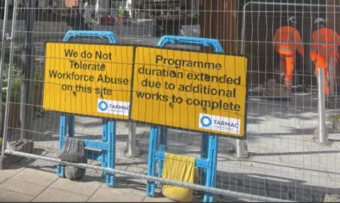 Yellow signs behind a metal fence on a building site. One says "We do not tolerate workforce abuse on this site" and the other days "Programme duration extended due to additional works to complete". Two workers in white hard hats and orange hi-vis overalls can be seen behind the signs. 