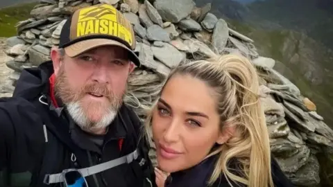Tim Edwards, wearing a baseball cap, poses for a photograph with his blonde-haired daughter Elle. They are standing at the top of Mount Snowdon. 