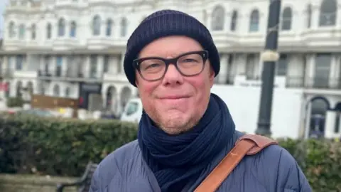 Josh McLaughlin/BBC Neill Morton smiles for the camera. He is stood in front of a row of hedges on Worthing promenade. He is wearing a navy beanie and scarf, black framed glasses and a brown satchel across his shoulder.
