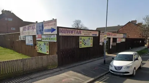 Google A Google Street View image of the shed supplier, with signs advertising the business on a fence outside. There is also a lawn and cars parked outside