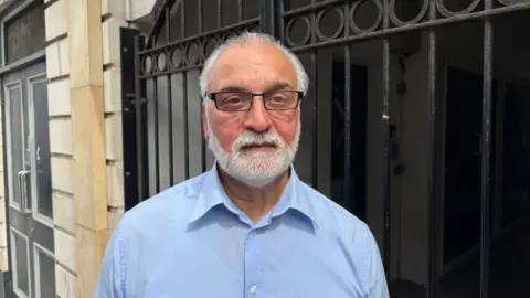 Martin Heath/BBC Raj Bains with short grey hair, a white beard and moustache, and rectangular glasses stands outside a building. He is wearing a light blue button-down shirt and is positioned in front of black metal railings and a recessed entrance. 
