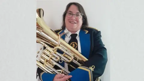 Rose Norman Rose in full black brass band suit with gold and blue trim. She is carrying a tuba.