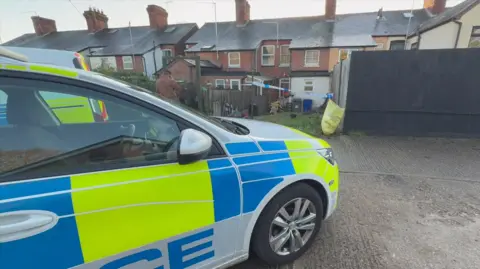 Jamie Niblock/BBC A police car is stood on concrete outside of a row of terraced houses. There is a blue and white police tape outside of one of the houses. 