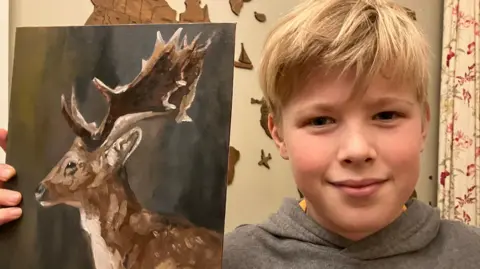 Reuben, with light hair and wearing a grey hoodie, smiles into the camera. He is holding a painting in his hands which depicts a large, brown deer.