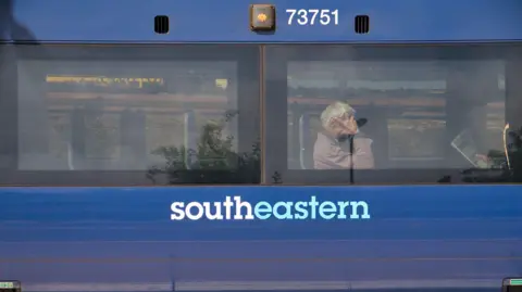 Getty Images The side of a blue Southeastern Railway carriage. Behind one of the two windows, commuters sit waiting for the train to continue its journey.