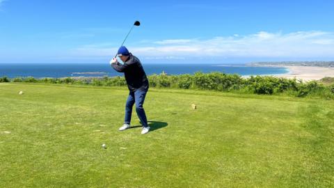 Pat raises his golf club above his head to tee off and behind him a long beach extends into the background