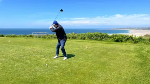 BBC Pat raises his golf club above his head to tee off and behind him a long beach extends into the background
