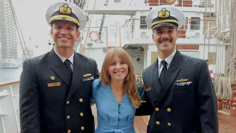 Two naval officers wearing smart uniforms and white military hats stand in the rain either side of a BBC reporter wearing a blue jumpsuit, on the wooden deck of a tallship, with ropes wound neatly behind them 