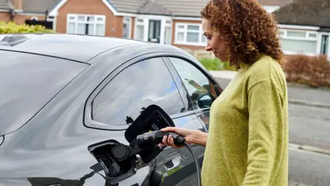 A woman with brown hair and a green top connects her electric car to a charging point