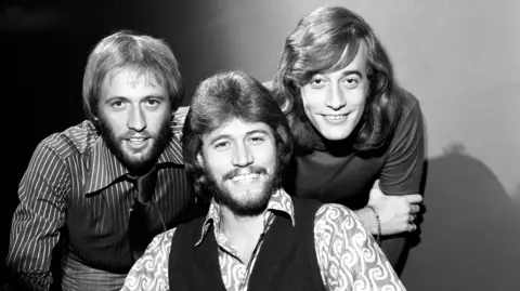 A black and white photo of the Bee Gees in the mid-70s. Maurice Gibb has short hair, a beard, with a striped shirt and trousers with a tie, Barry Gibb has longer hair and a beard, and wears a patterned shirt and waistcoat, and Robin Gibb has no beard and long hair, and is wearing a polo neck. They are backstage at Top of the Pops.