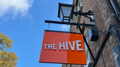 The Hive sign; it's bright orange with white letters reading 'The Hive'. It's hung off the side of the building like a pub sign, with a lantern above it. The photo is shot looking upwards, so the sign is pictured against a bright blue sky with some cloud. 