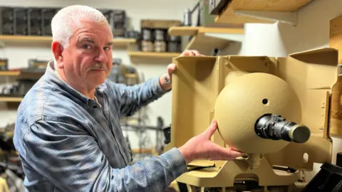 BBC Grame Delanoe stands with two hands on a machine gun in an armoured turret which has been restored to its original condition