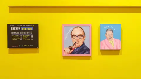 Tom Bird Canvas paintings hung on a yellow gallery walls depicting Ceefax and comedians Morecambe and Wise. Morecambe is holding a pipe to his mouth and Wise smiling broadly in a pink suit